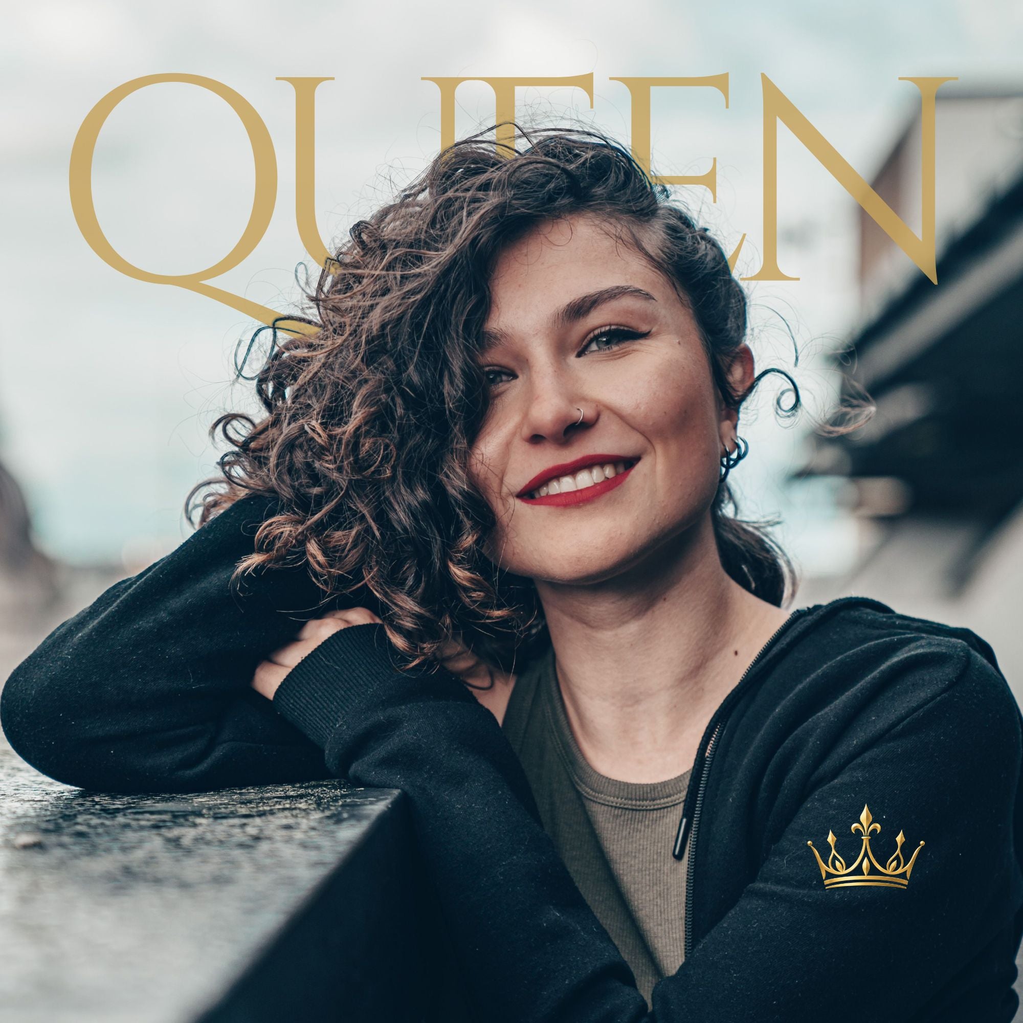 A smiling woman with curly brown hair leans on a stone wall, wearing a black zip-up hoodie with a small gold crown logo on the left chest. Large gold text reading 'QUEEN' overlays the top of the image.
