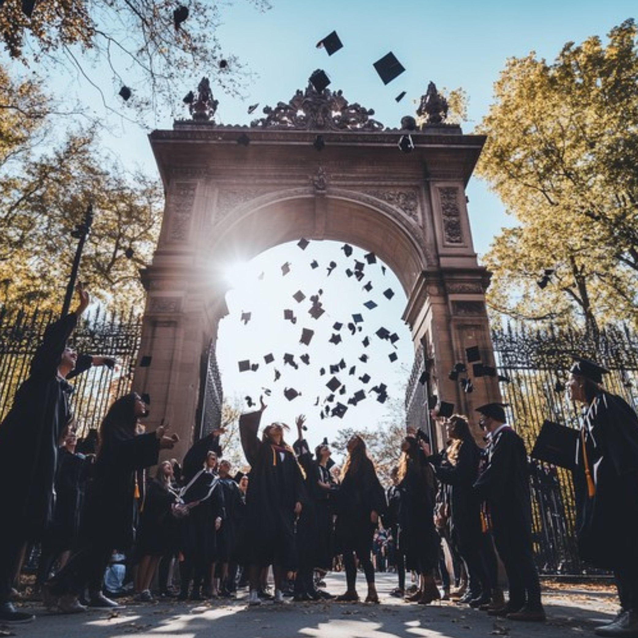 Graduates throwing hats in the air in front of an archway with trees and sunlight.