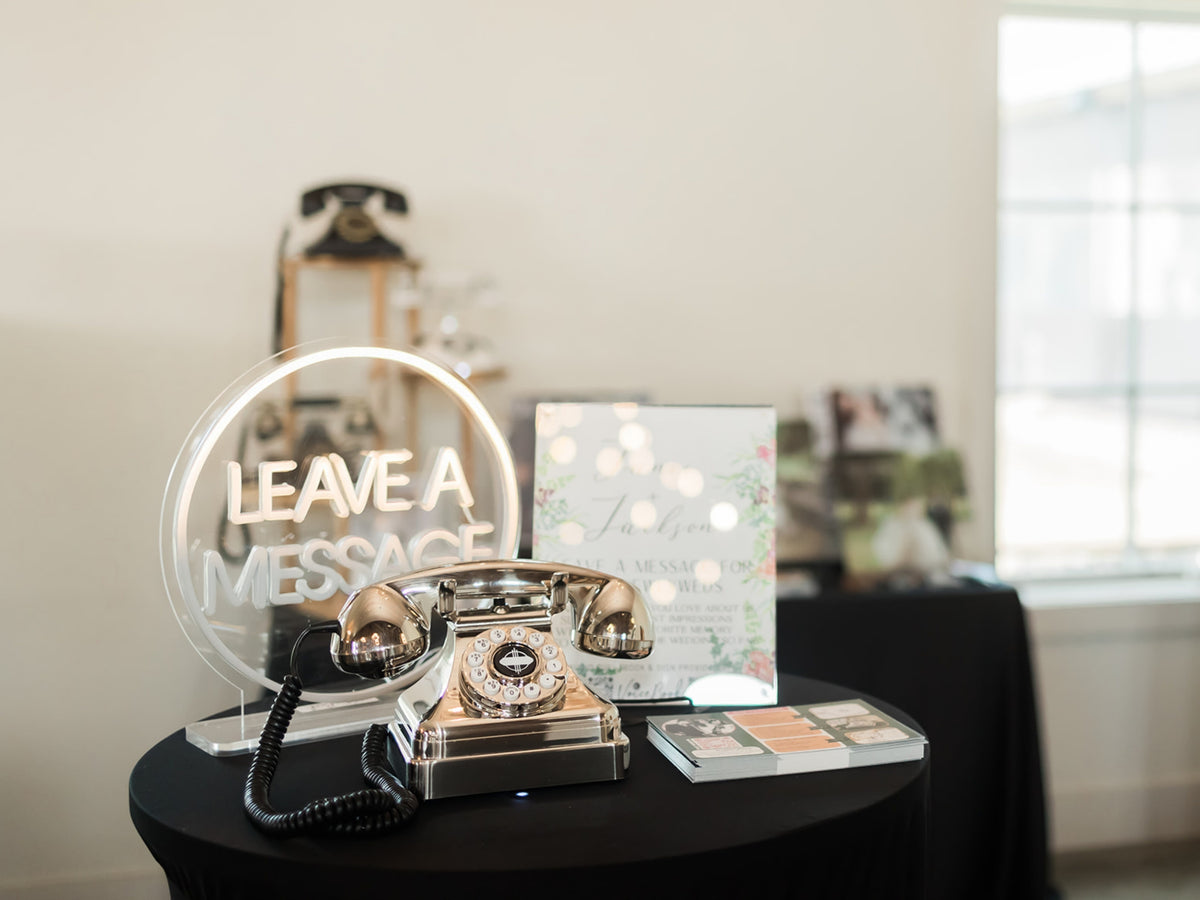 A vintage-style chrome rotary telephone sits on a black table beside a clear acrylic sign reading 'LEAVE A MESSAGE' and a floral invitation card in a softly lit indoor setting.