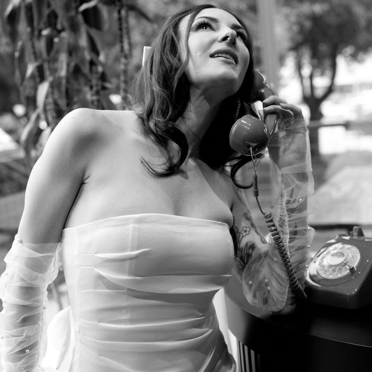 Black and white photograph of a woman in a strapless white dress holding a vintage rotary telephone receiver, with a matching rotary phone visible on a table beside her.