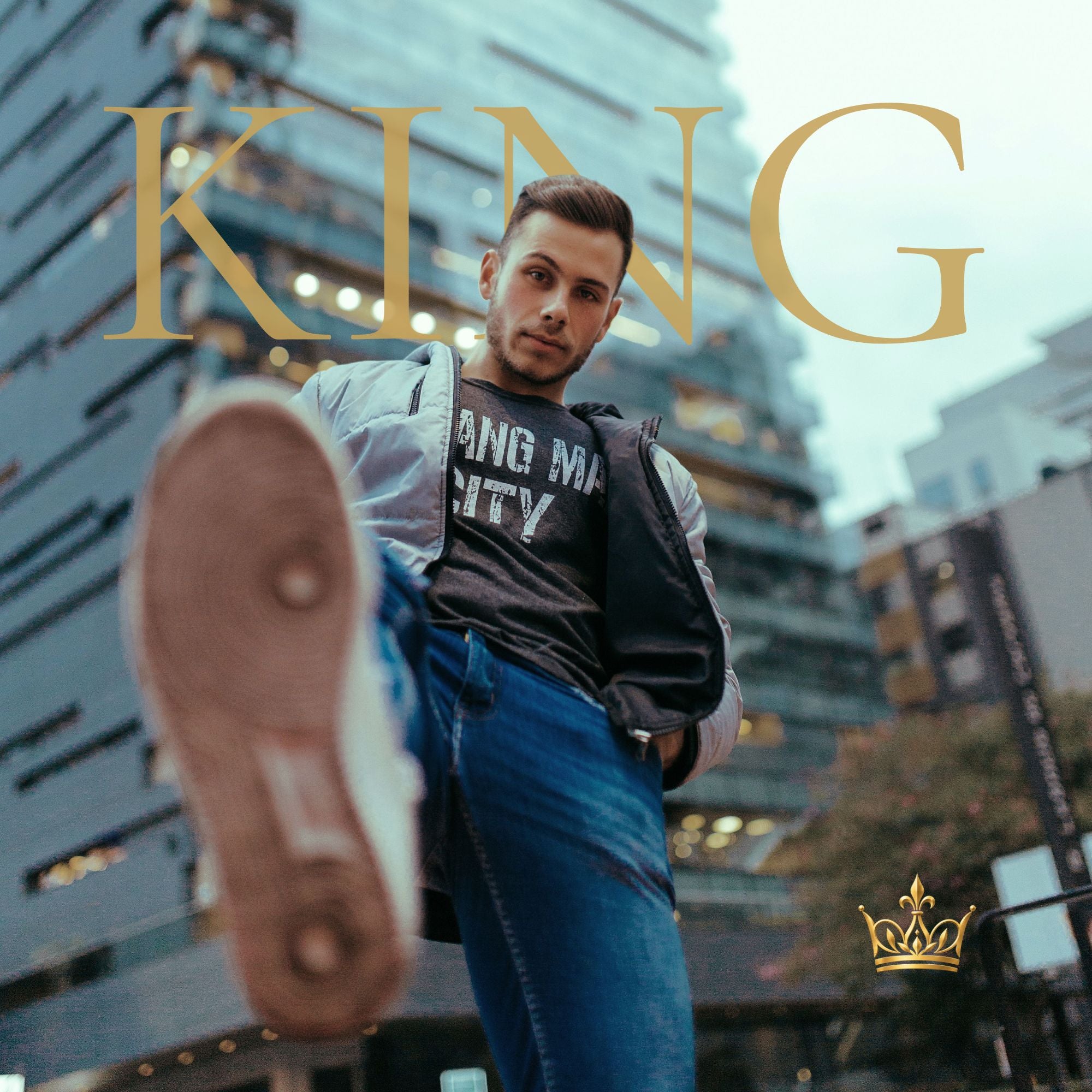 A man in a city setting wearing a gray graphic t-shirt, black jacket, and blue jeans, with his shoe raised in the foreground. Large gold text reads 'KING' across the top, and a small gold crown logo appears in the bottom right corner.