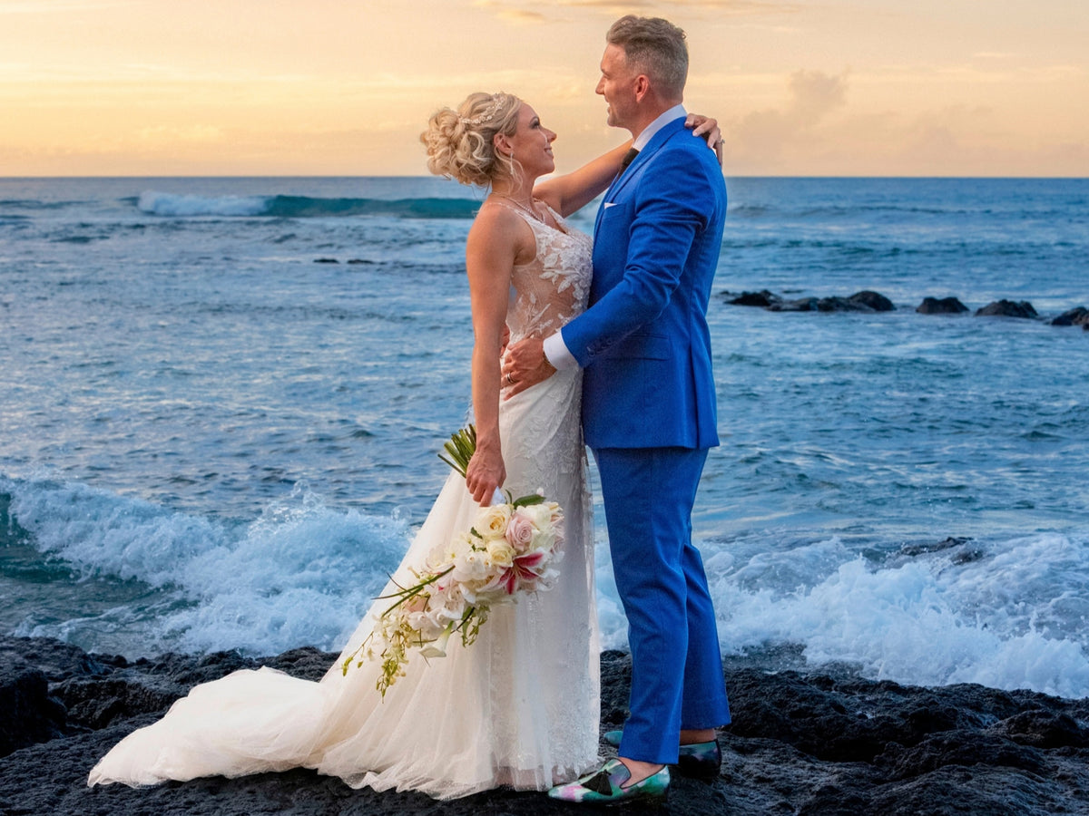 A bride in a white lace wedding gown holding a floral bouquet and a groom in a bright blue suit embracing on a rocky ocean shore at sunset.