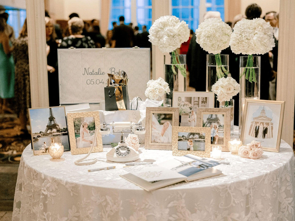 A round white table covered with a lace tablecloth displays a custom bridal registry setup. The table is decorated with multiple framed wedding photographs, a vintage-style white rotary phone, a small white typewriter, candles, and white floral arrangements in tall glass vases.