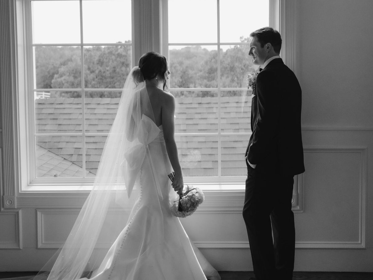 Black and white wedding photograph showing a bride in a sleeveless white gown with a long veil holding a bouquet, standing beside a groom in a dark suit in front of a large window.