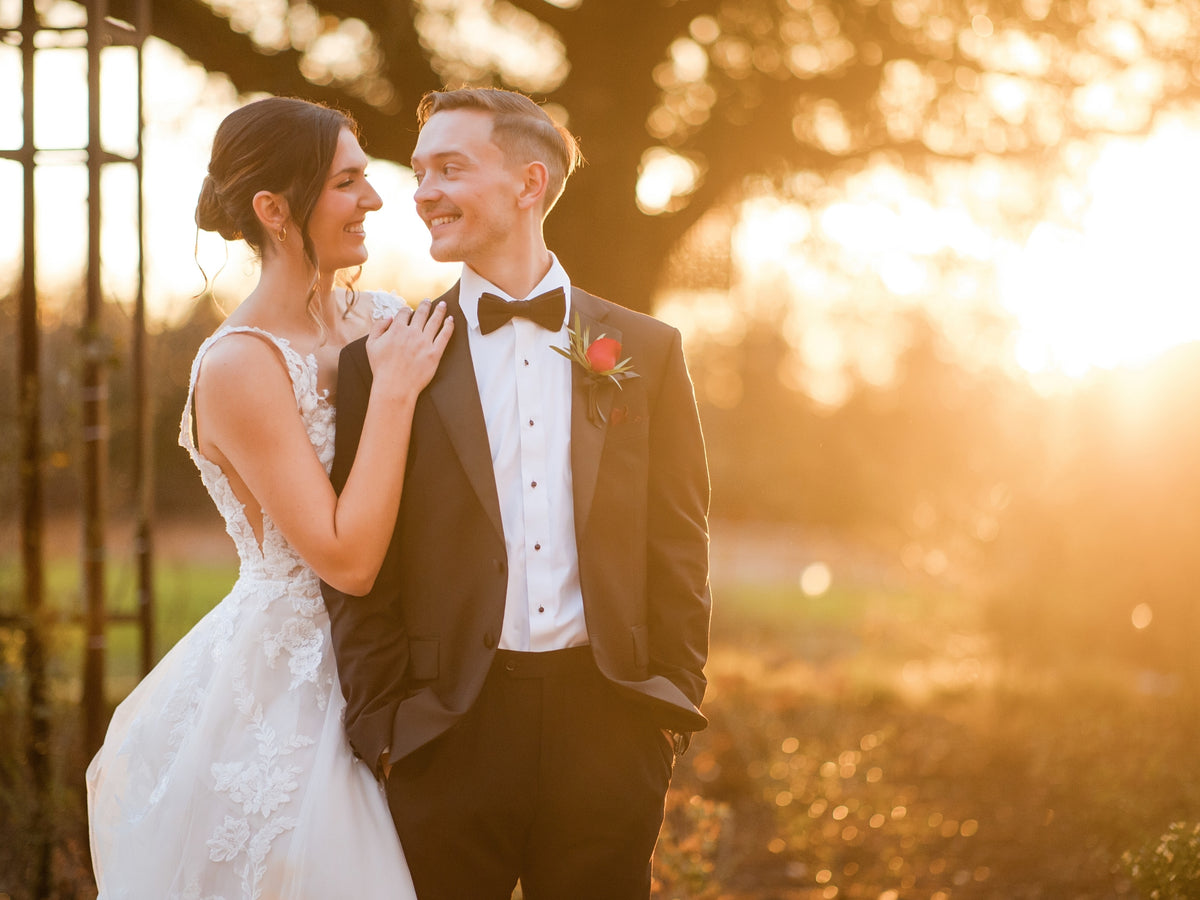 A bride in a white lace wedding gown and a groom in a black tuxedo with a white shirt and black bow tie stand close together outdoors at sunset. The background is softly blurred with warm golden light filtering through trees.